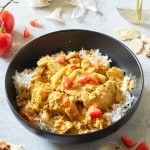 A black bowl filled with white rice topped with a creamy chicken curry garnished with chopped tomatoes. Surrounding the bowl are cherry tomatoes, garlic cloves, fresh ginger, a spoon with curry sauce, and pieces of naan. A glass jar with yellow flowers is in the background.