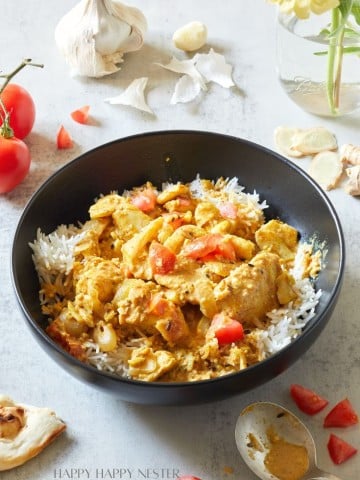 A black bowl filled with white rice topped with a creamy chicken curry garnished with chopped tomatoes. Surrounding the bowl are cherry tomatoes, garlic cloves, fresh ginger, a spoon with curry sauce, and pieces of naan. A glass jar with yellow flowers is in the background.