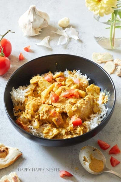 A black bowl filled with white rice topped with a creamy chicken curry garnished with chopped tomatoes. Surrounding the bowl are cherry tomatoes, garlic cloves, fresh ginger, a spoon with curry sauce, and pieces of naan. A glass jar with yellow flowers is in the background.