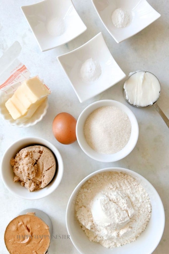 Overhead view of baking ingredients arranged on a counter: small bowls of salt, baking powder, and baking soda; a half stick of butter; one egg; bowls of white sugar, brown sugar, and flour; and a cup of shortening. .