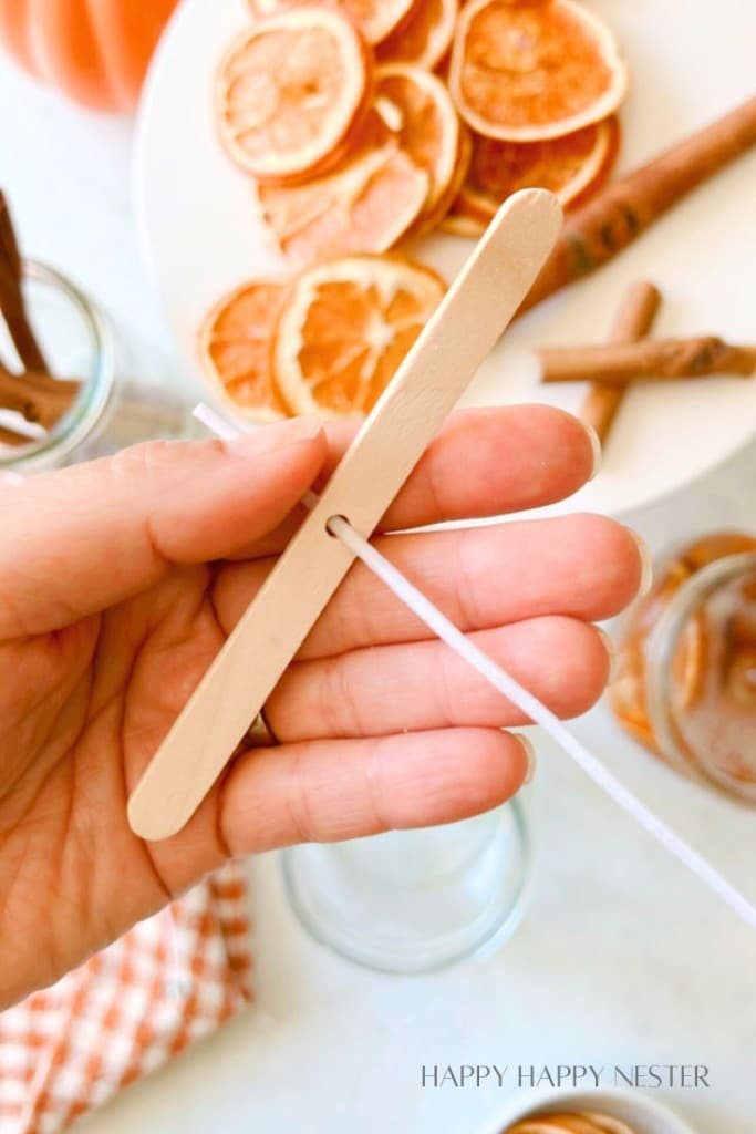 A hand holding a wooden stick, threading a string through a small hole in the center. In the background, a white plate with dried orange slices and a couple of cinnamon sticks are visible. The edge of a checkered cloth can be seen at the bottom.