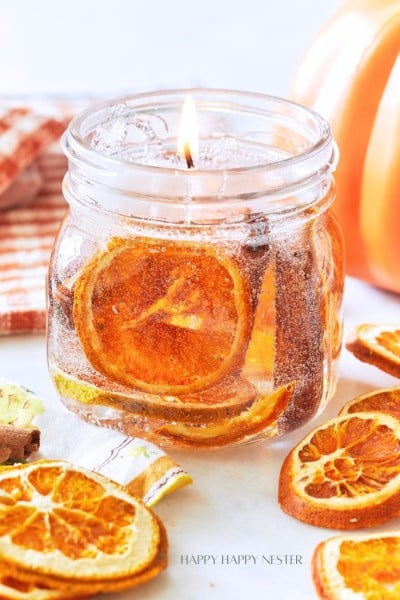 A lit candle in a small glass jar, adorned with dried orange slices and cinnamon sticks, sits on a surface alongside additional dried orange slices. A striped cloth napkin and a partially visible pumpkin decor can be seen in the background.