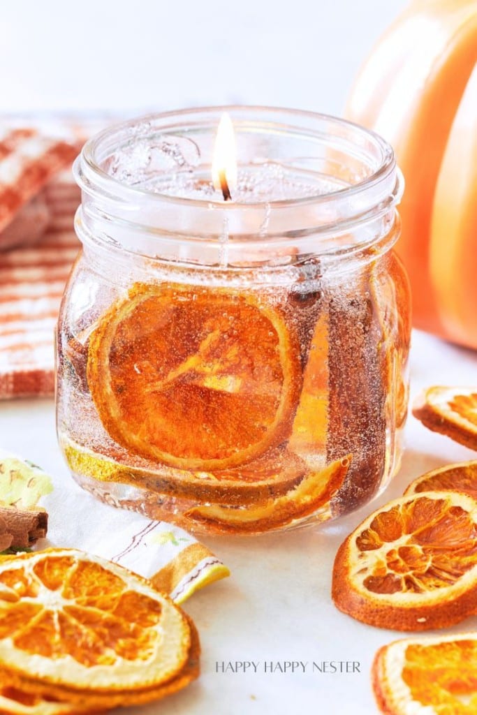 A lit candle in a small glass jar, adorned with dried orange slices and cinnamon sticks, sits on a surface alongside additional dried orange slices. A striped cloth napkin and a partially visible pumpkin decor can be seen in the background.