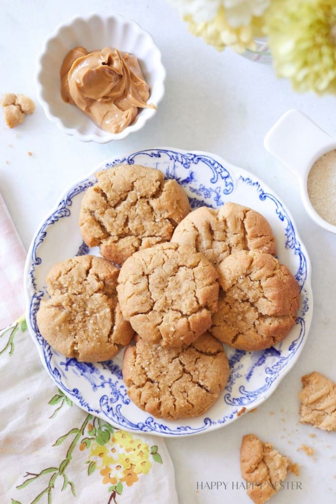 A blue and white plate holds six peanut butter cookies, some with cracked tops, on a light background. Nearby are a small bowl of peanut butter, a bowl of sugar, a floral cloth, and a bouquet of flowers. Some cookie crumbs are scattered around.