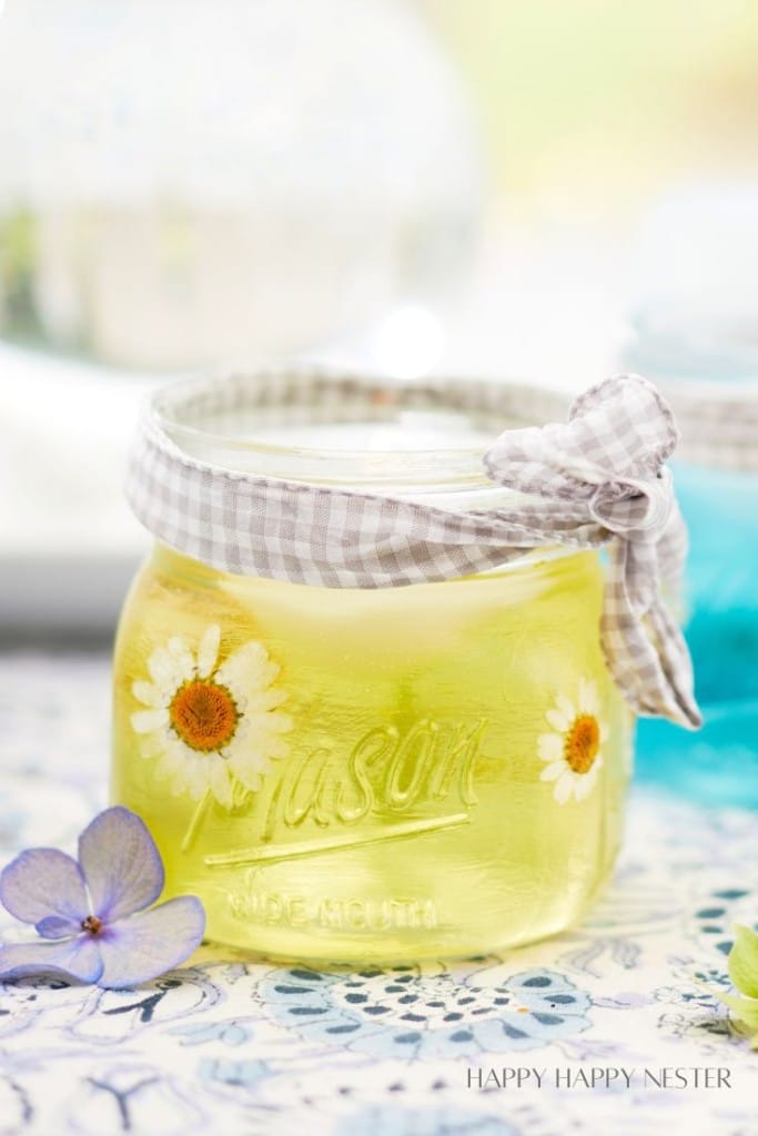 A Mason jar filled with a yellowish liquid, decorated with small white daisies. The jar is covered with a gingham cloth tied with a bow. The jar is placed on a floral-patterned surface, and a purple hydrangea flower is in the foreground.