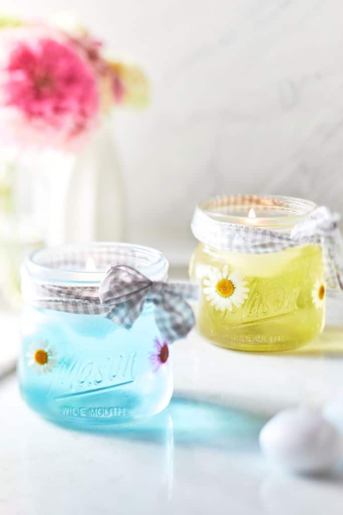 Two mason jar candles, one with blue wax and the other with pale yellow wax, are decorated with daisy flowers and tied with checkered ribbons. A blurred bouquet of pink flowers is in the background.