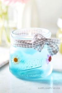 A blue candle is set in a Mason jar adorned with pressed flowers and tied with a checkered gray and white ribbon. The candle is lit, and the background is softly blurred, creating a cozy ambiance. The words "HAPPY HAPPY NESTER" are visible in the bottom right corner.