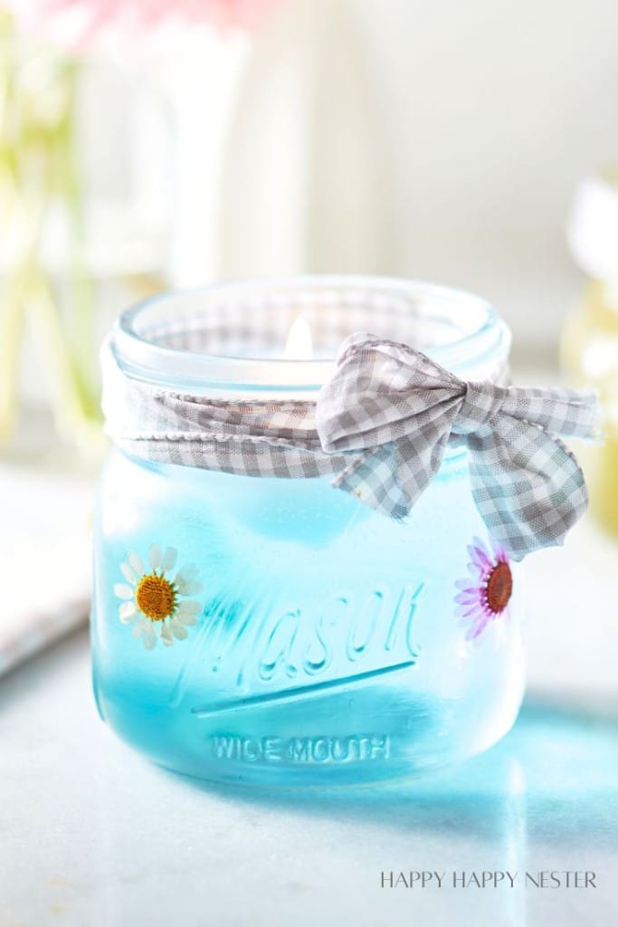 A blue candle is set in a Mason jar adorned with pressed flowers and tied with a checkered gray and white ribbon. The candle is lit, and the background is softly blurred, creating a cozy ambiance. The words "HAPPY HAPPY NESTER" are visible in the bottom right corner.