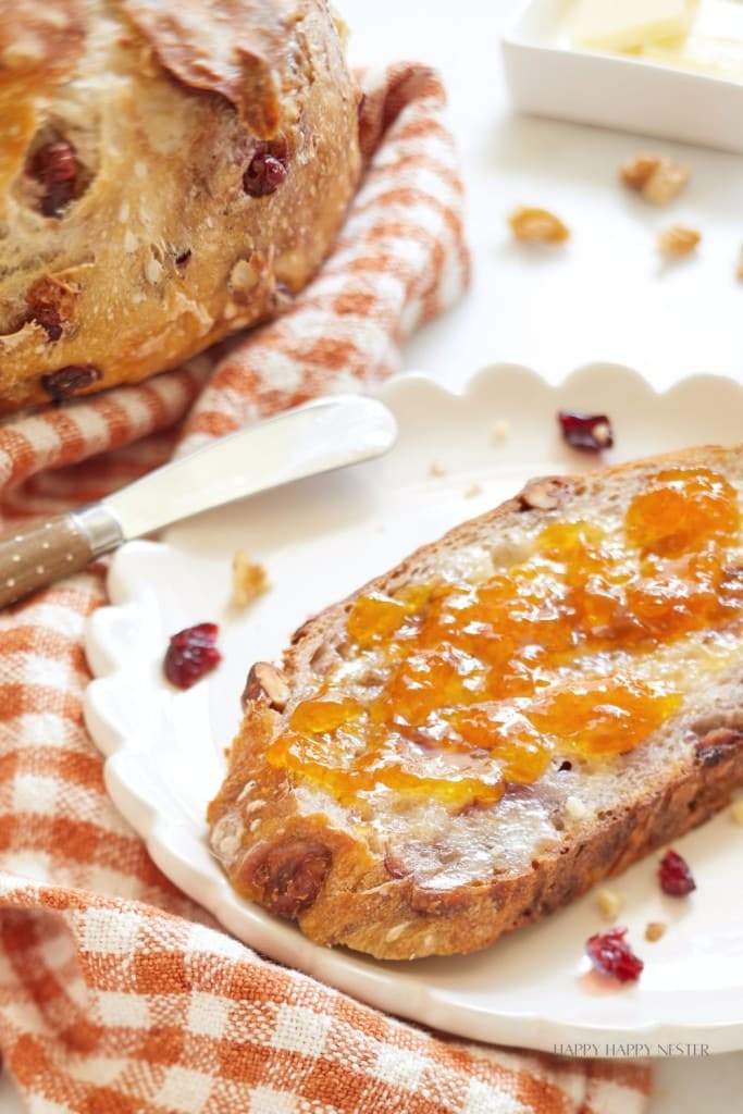 A close-up of a slice of rustic bread spread with orange marmalade on a white scalloped plate. Next to it is a whole loaf of bread and a butter knife resting on a checked cloth napkin. Some dried cranberries and walnuts are scattered around.