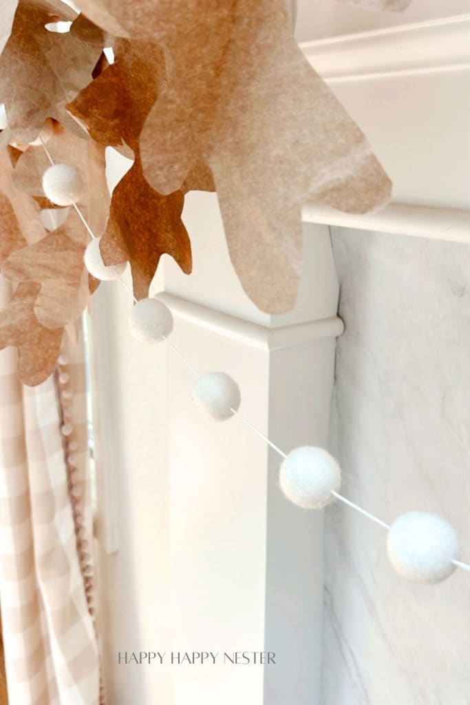 A close-up shot shows a decorative garland draped across a white mantel. The garland consists of large brown paper leaves and white spherical ornaments. The background includes a marble surface and a partially visible checkered curtain.