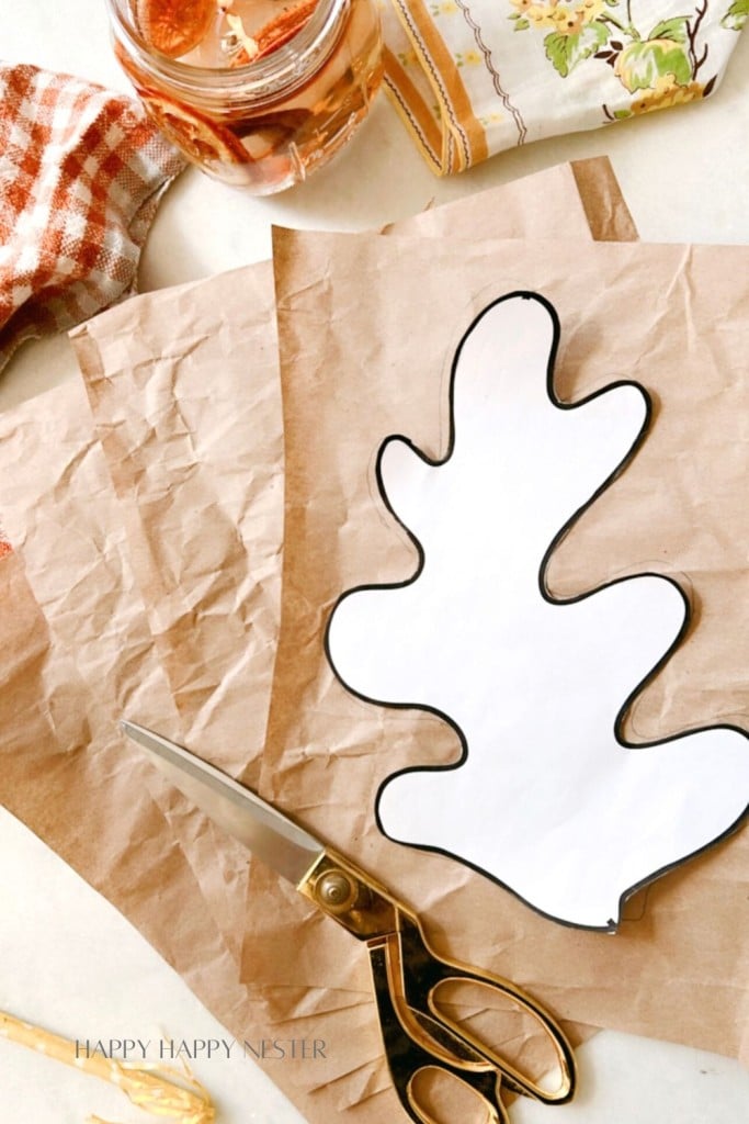 An overhead view of a crafting scene with crumpled brown paper, a white paper leaf cutout with a black outline, a pair of gold-handled scissors, and a red and white checkered cloth. A jar and a patterned cloth are also visible in the background.