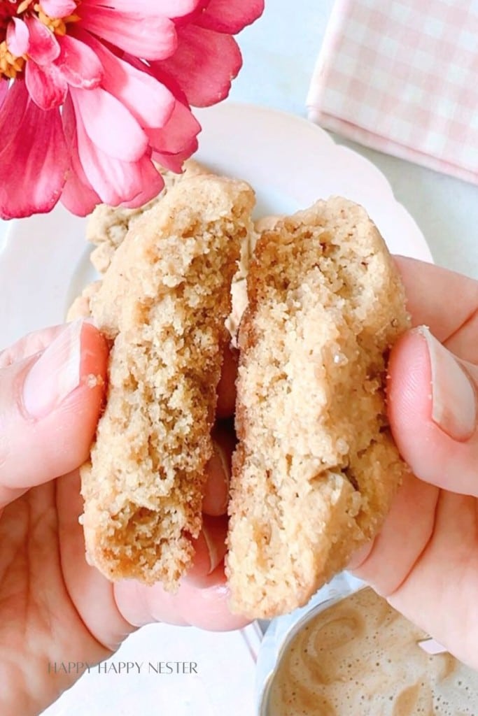 A close-up of hands breaking apart a crumbly homemade cookie with a pink flower in the background. A plate and a mug of coffee are partially visible. The words "Happy Happy Nester" are subtly printed at the bottom.