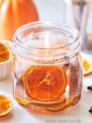 A mason jar candle with a lit flame contains dried orange slices and spices, creating a rustic and cozy ambiance. The background is softly blurred with a pumpkin and additional dried orange slices surrounding the jar.