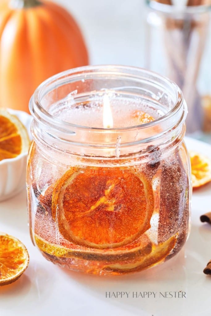 A mason jar candle with a lit flame contains dried orange slices and spices, creating a rustic and cozy ambiance. The background is softly blurred with a pumpkin and additional dried orange slices surrounding the jar.