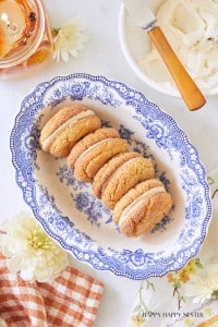 A decorative blue and white plate holds four sandwiched cookies with a creamy filling. The plate is set on a marble surface, surrounded by a dish of more filling, a white flower, an orange checked cloth, and a glass of amber-colored drink.