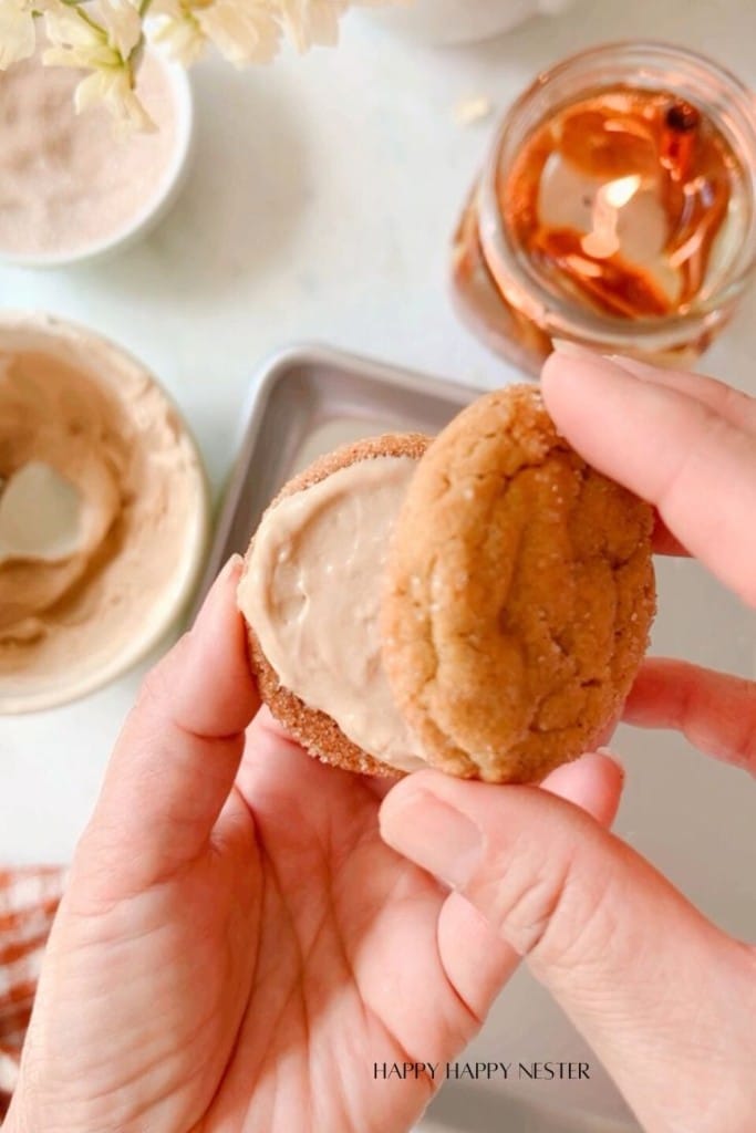 A person holds two cookies sandwiched together with creamy frosting. A bowl with remaining frosting, a tray, and a jar with a lit candle are in the background on a light-colored surface. White flowers and a brown checkered cloth are partially visible.