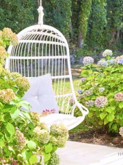 A white hanging chair with a grey cushion is set in a lush garden. The chair is surrounded by blooming hydrangea bushes in shades of pink and blue, with greenery and a stone pathway completing the tranquil outdoor scene.