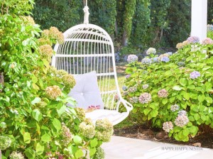 A white hanging chair with a grey cushion is set in a lush garden. The chair is surrounded by blooming hydrangea bushes in shades of pink and blue, with greenery and a stone pathway completing the tranquil outdoor scene.