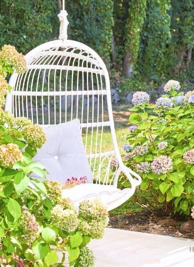 A white hanging chair with a grey cushion is set in a lush garden. The chair is surrounded by blooming hydrangea bushes in shades of pink and blue, with greenery and a stone pathway completing the tranquil outdoor scene.