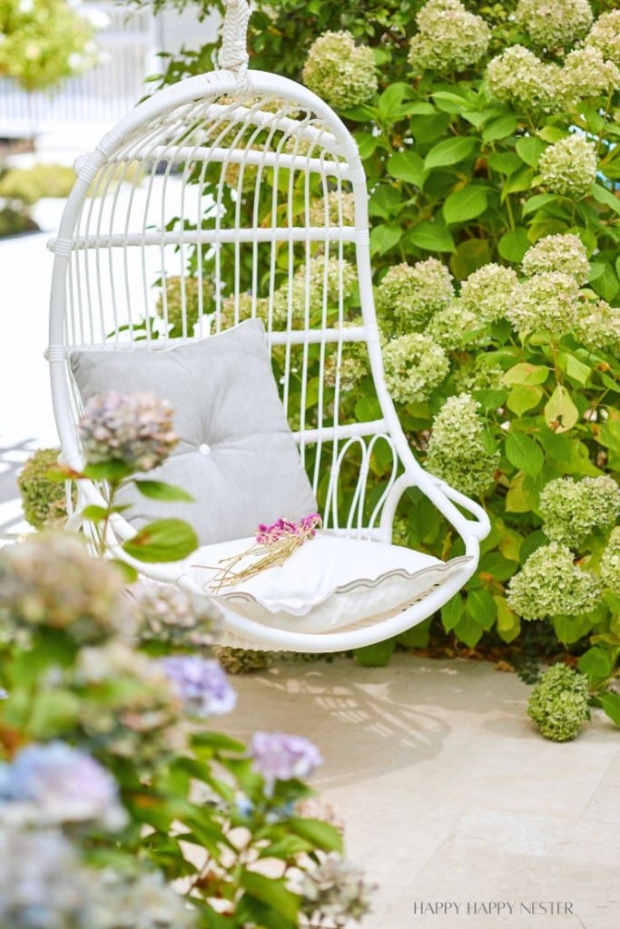 White rattan hanging chair with a grey cushion, adorned with a small bouquet of flowers, positioned in a garden. The background features blooming hydrangea bushes with light greenish-white flowers. Sunlight illuminates the scene, creating a serene outdoor setting.