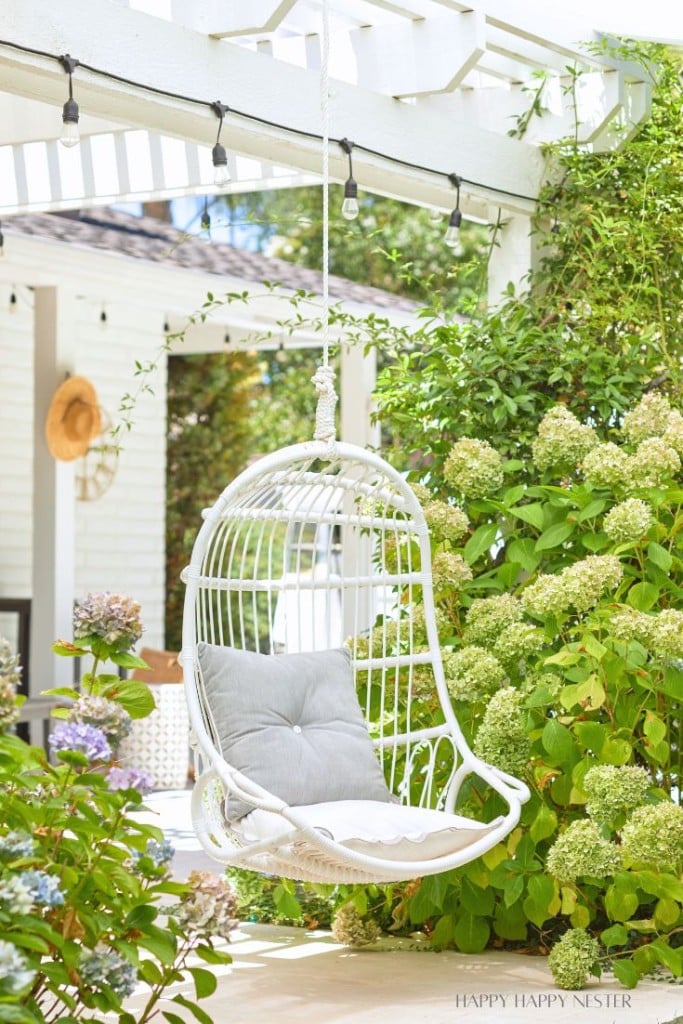 A white wicker hanging chair with gray cushions is suspended from a pergola in a lush garden. The pergola is adorned with hanging string lights, and there are green bushes and blooming flowers around. A hat hangs on the white wall in the background.