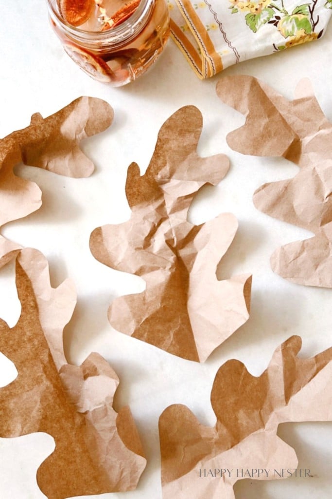 Crumpled paper leaves are arranged artistically on a white surface with a mason jar containing dried orange slices and a folded patterned napkin nearby.