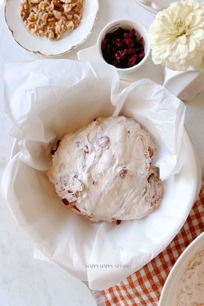 A bowl of bread dough with cranberries sits on a table covered with a piece of parchment paper. Surrounding the bowl are small bowls containing chopped nuts and dried cranberries. A beige and white checkered towel is partially visible under the bowl.