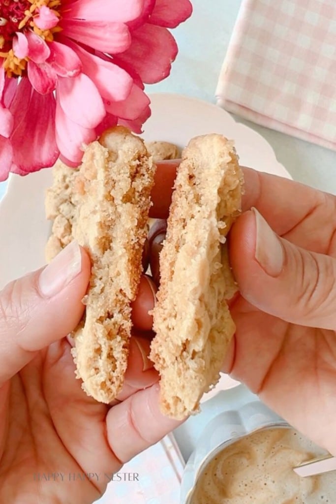 Two hands holding a halved cookie with a chunky texture, revealing its slightly crumbly interior. In the background, there is a pink flower and a light-colored saucer. A cup of frothy coffee and a small checkered cloth are also partially visible.