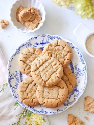 A plate of peanut butter cookies arranged on a decorative blue and white plate. A small bowl of peanut butter, a jug with brown sugar, and cookie crumbs are beside the plate. A pink and green floral towel and yellow flowers partially frame the scene.