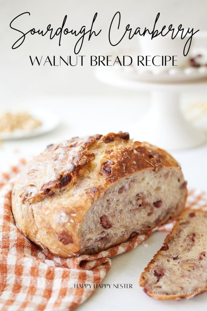 A loaf of sourdough cranberry walnut bread sits on an orange and white checkered cloth, with one slice cut from it. In the background, there are walnuts and a cake stand. The text reads "Sourdough Cranberry Walnut Bread Recipe.