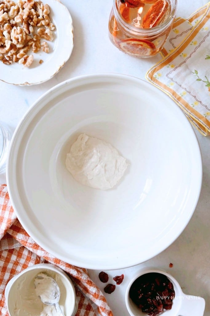 A large white mixing bowl containing a lump of dough is centered on a white surface. Surrounding the bowl are various ingredients in dishes, including walnuts, a jar of liquid, and small amounts of other items. An orange-checked cloth is laid beside the bowl.