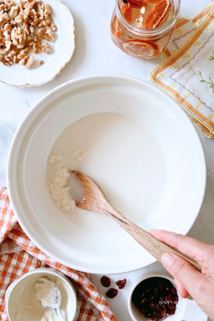 A top view of a wooden spoon stirring a white mixture in a large white mixing bowl. Surrounding the bowl on a light counter are chopped walnuts on a plate, a jar of preserved lemons, a checked cloth, and small bowls containing ingredients.