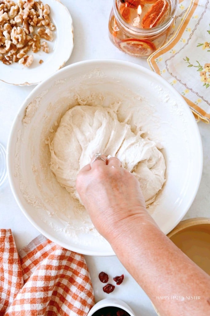 A person kneads dough in a white bowl. Surrounding the bowl are a plate of walnuts, a jar with orange slices, a checkered cloth, and some cranberries. The scene takes place on a white surface.