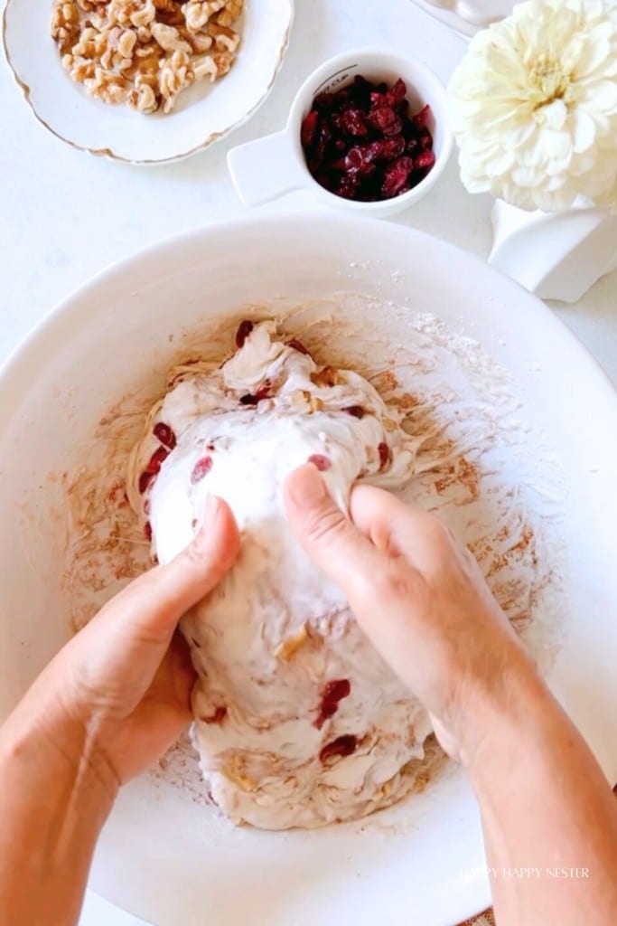 Two hands are kneading dough with visible pieces of dried cranberries and nuts. Nearby, there are bowls containing walnuts and dried cranberries, and a white flower in the background. The scene suggests preparation of a homemade baked good.