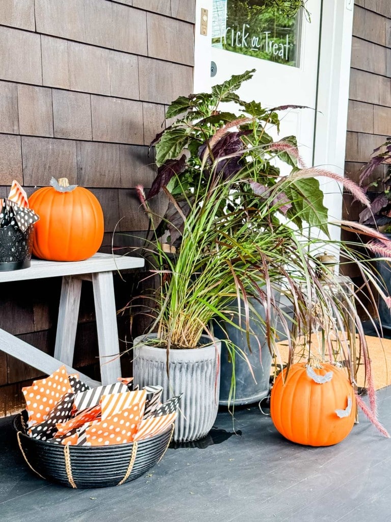 A festive porch decorated for Halloween with pumpkins, a basket of orange and black patterned treat bags on a bench, several potted plants, and a charming DIY paper garland. A door in the background has a "Trick or Treat" sign, and one pumpkin is carved into a jack-o'-lantern.