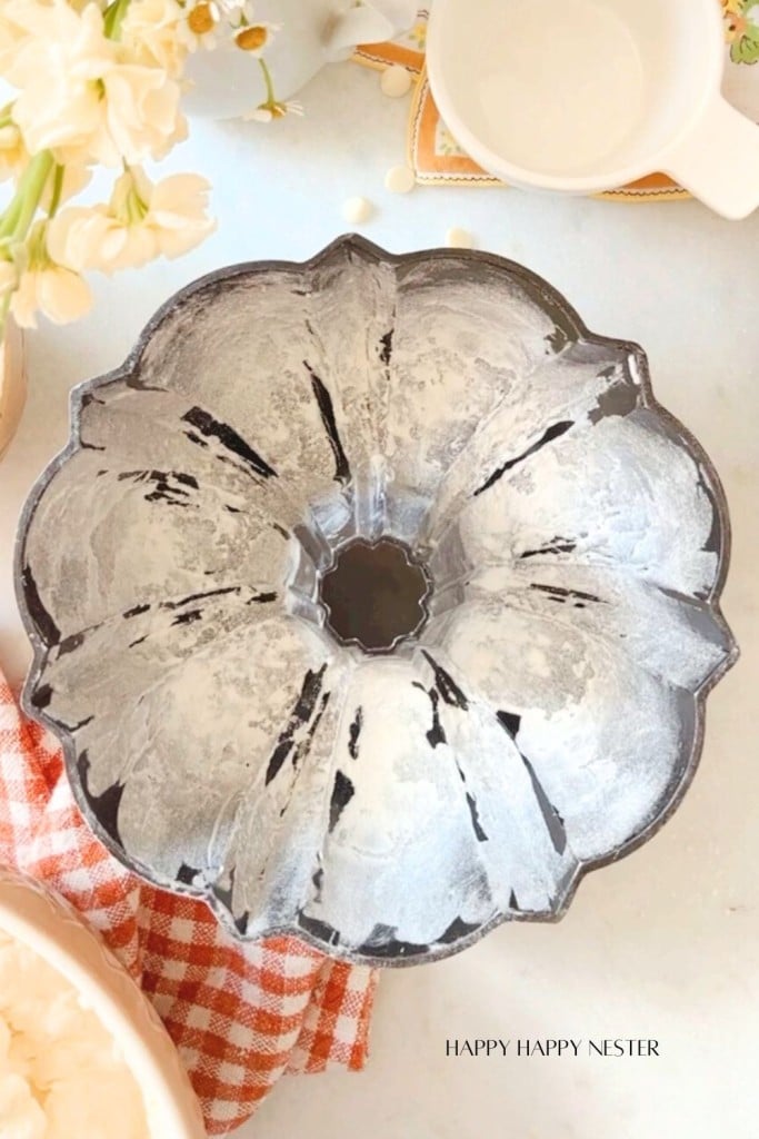 A fluted Bundt cake pan with flour coating is placed on a kitchen countertop. Nearby, there are white flowers in a vase, a white mug, and a checkered red and white cloth. In the bottom right corner, the text "Happy Happy Nester" can be seen.