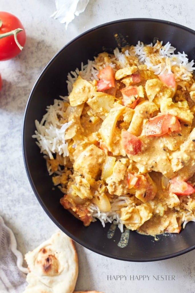 A savory dish consisting of rice topped with a creamy curry featuring chunks of chicken and garnished with diced tomatoes is served in a black bowl. The dish is accompanied by a side of naan bread. A tomato and napkin are visible nearby on the tabletop.