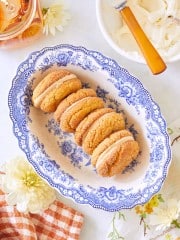 A blue and white floral platter holds six cookies with cream filling, stacked neatly in two rows. Nearby, there is a jar of honey, a bowl with a knife, and a checkered cloth napkin. White and yellow flowers add a decorative touch to the arrangement.