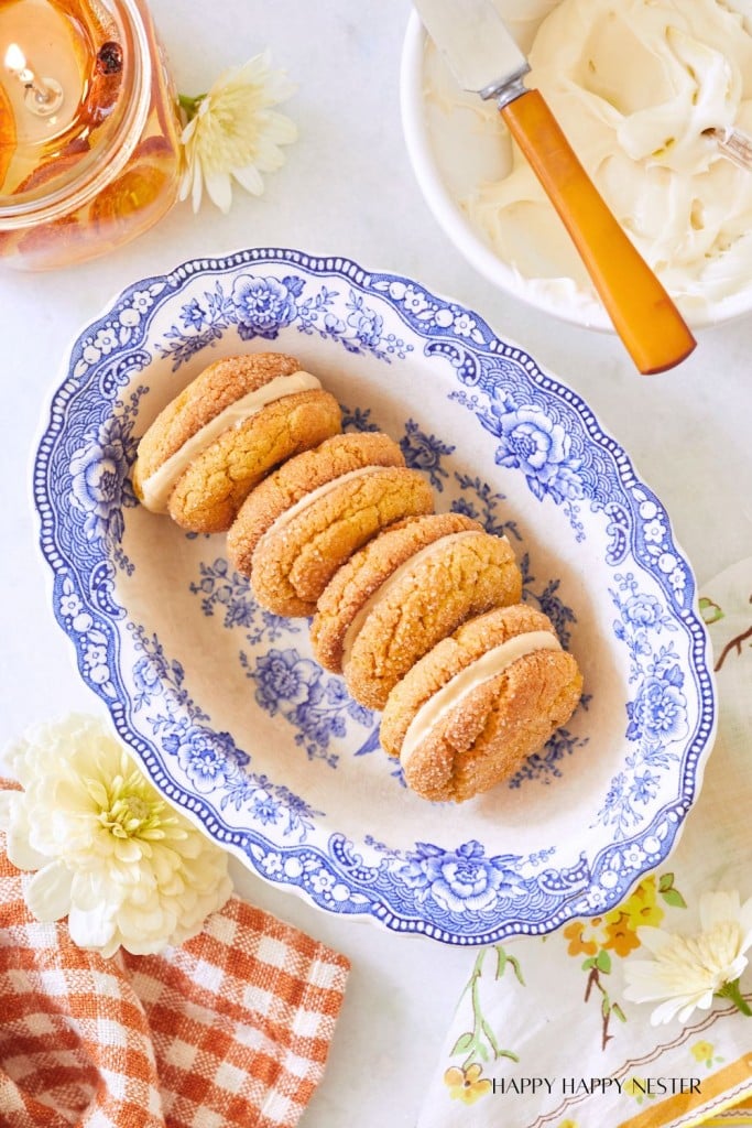 A blue and white floral platter holds six cookies with cream filling, stacked neatly in two rows. Nearby, there is a jar of honey, a bowl with a knife, and a checkered cloth napkin. White and yellow flowers add a decorative touch to the arrangement.