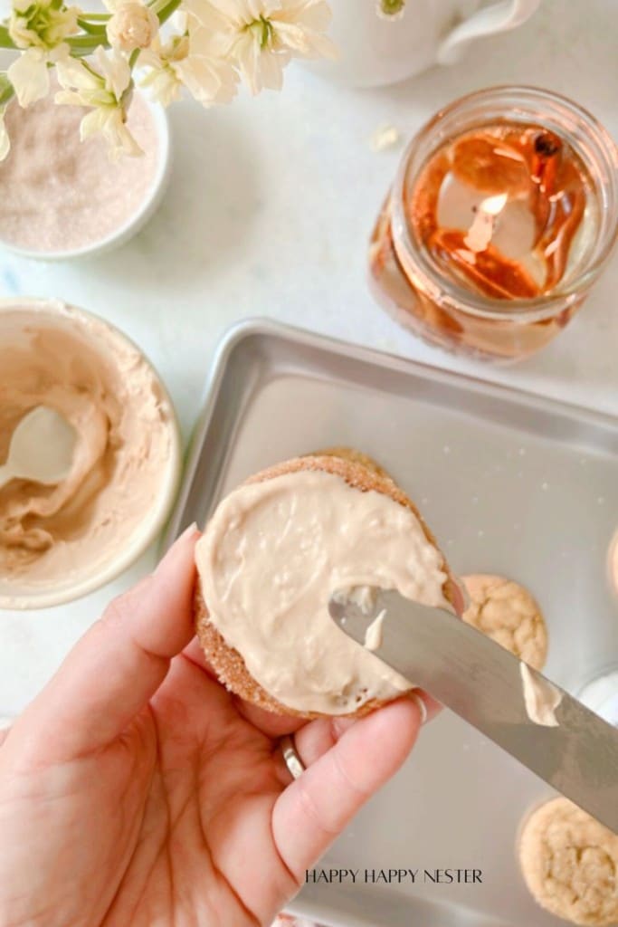 A hand is spreading light brown frosting on a cookie using a knife. Nearby, there are bowls of frosting and sugar, a metal tray with cookies, and a mason jar filled with a brown liquid. Flowers and a teapot are in the background. Text at the bottom reads, "HAPPY HAPPY NESTER.