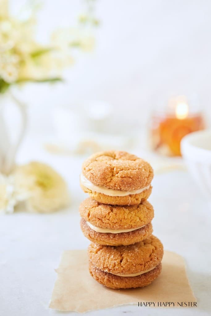 A stack of four peanut butter sandwich cookies sits on a piece of parchment paper on a white surface. The background is softly blurred, featuring a white ceramic pitcher with flowers, a lit candle, and a bowl. The image has a serene and cozy ambiance.