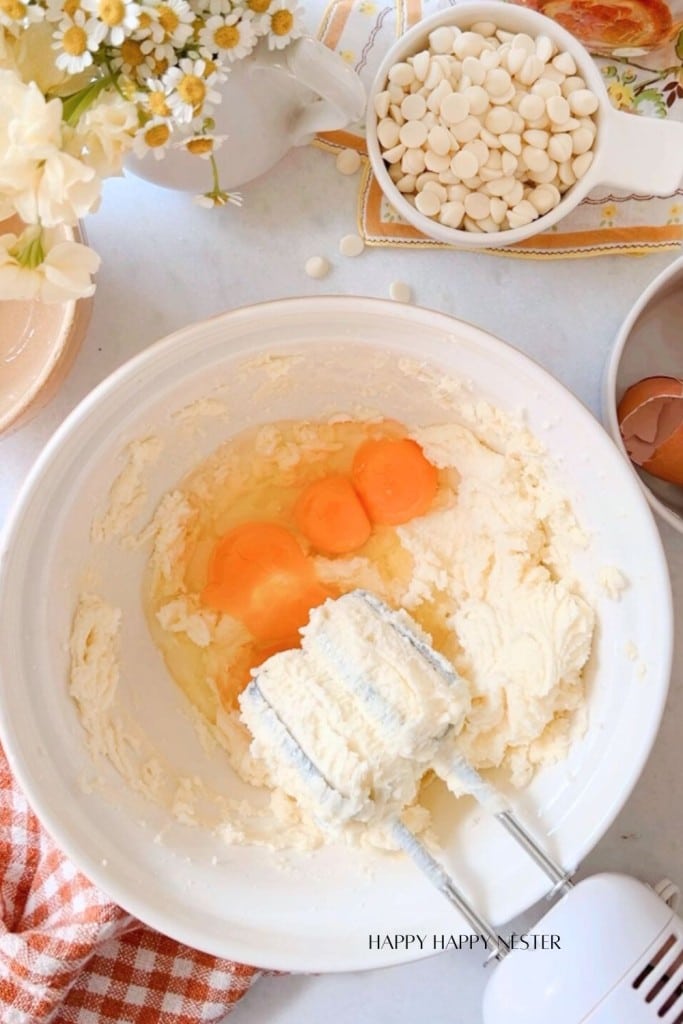 A large white mixing bowl holds a light-colored batter with two cracked eggs on top. A hand mixer with beaters is in the bowl, surrounded by baking ingredients, a bowl of white chocolate chips, cracked eggshells, and flowers. The text "HAPPY HAPPY NESTER" is at the bottom.