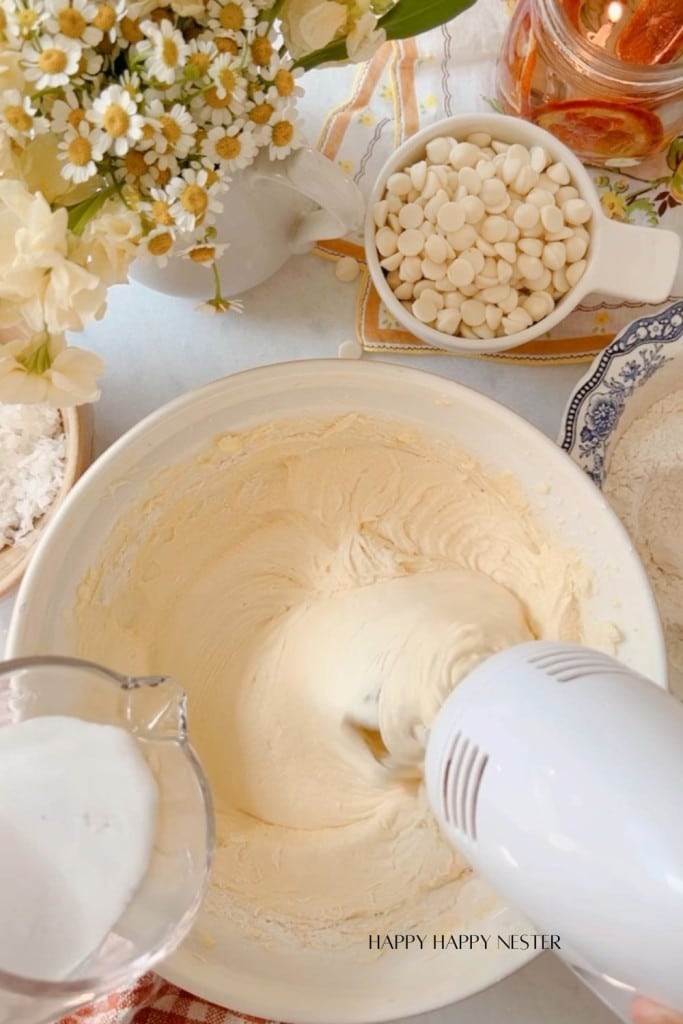 A hand mixer is blending batter in a mixing bowl on a kitchen countertop. Surrounding the bowl are ingredients for baking, including a measuring cup with sugar, a bowl of white chocolate chips, a jar of oil, a pitcher with flowers, and a dish with flour.