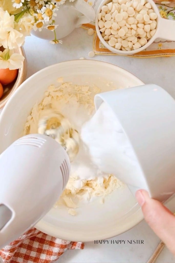 An electric mixer blending a white creamy mixture in a bowl. A hand is pouring more mixture from a white cup. On the table are a small bowl with white chocolate chips, white flowers, and a checkered cloth. The text "Happy Happy Nester" is at the bottom.