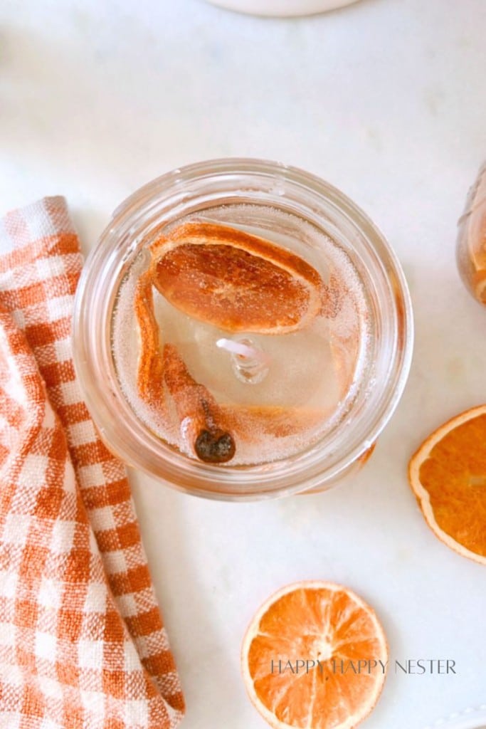 A mason jar filled with a drink, garnished with a dried citrus slice and a cinnamon stick. Next to it, there's a checkered orange and white napkin and two additional dried citrus slices on a flat surface.