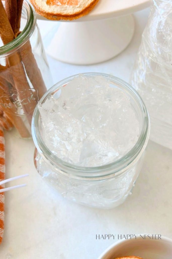 A clear glass jar filled with transparent shards of what appears to be homemade gel or jelly sits on a white surface. Next to the jar is a checkered cloth, some cinnamon sticks in another jar, and dried orange slices.