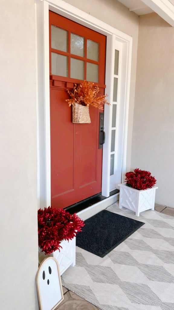 A red front door adorned with a fall-themed wreath featuring orange leaves, flanked by small rectangular windows. Two white planters with vibrant red flowers sit on either side of the entrance. For inspiring fall decor ideas, a black doormat and a white ghost decoration lie on the doorstep.