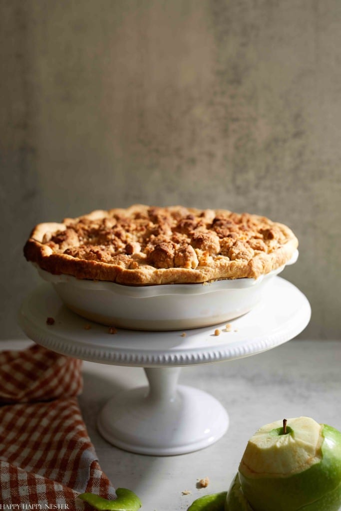 A golden-brown apple crumble pie sits on a white pedestal cake stand, embodying the essence of autumn recipes. The pie crust is flaky with a crumbly top. Next to the stand is a checkered red napkin and a partially peeled green apple with apple peels scattered beside it. Neutral background.