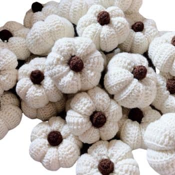 A cluster of crocheted white pumpkins with brown stems, piled together. The pumpkins have a textured appearance, emphasizing the intricate knit patterns. The overall tone is cozy and craft-inspired.