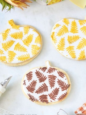 A close-up of four cookies shaped like pumpkins, each decorated with intricate fern leaf designs. Three have yellow and white icing, and one has brown and white icing. A piping bag is visible on the side, and there's a sunny, autumn-themed decoration nearby.